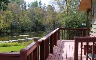 A red wooden deck attached to a house overlooking a calm pond surrounded by trees.