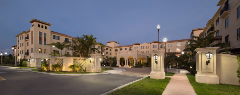 Exterior view of a large, multi-story senior living facility building at dusk with well-lit entrance, palm trees, and landscaped grounds.