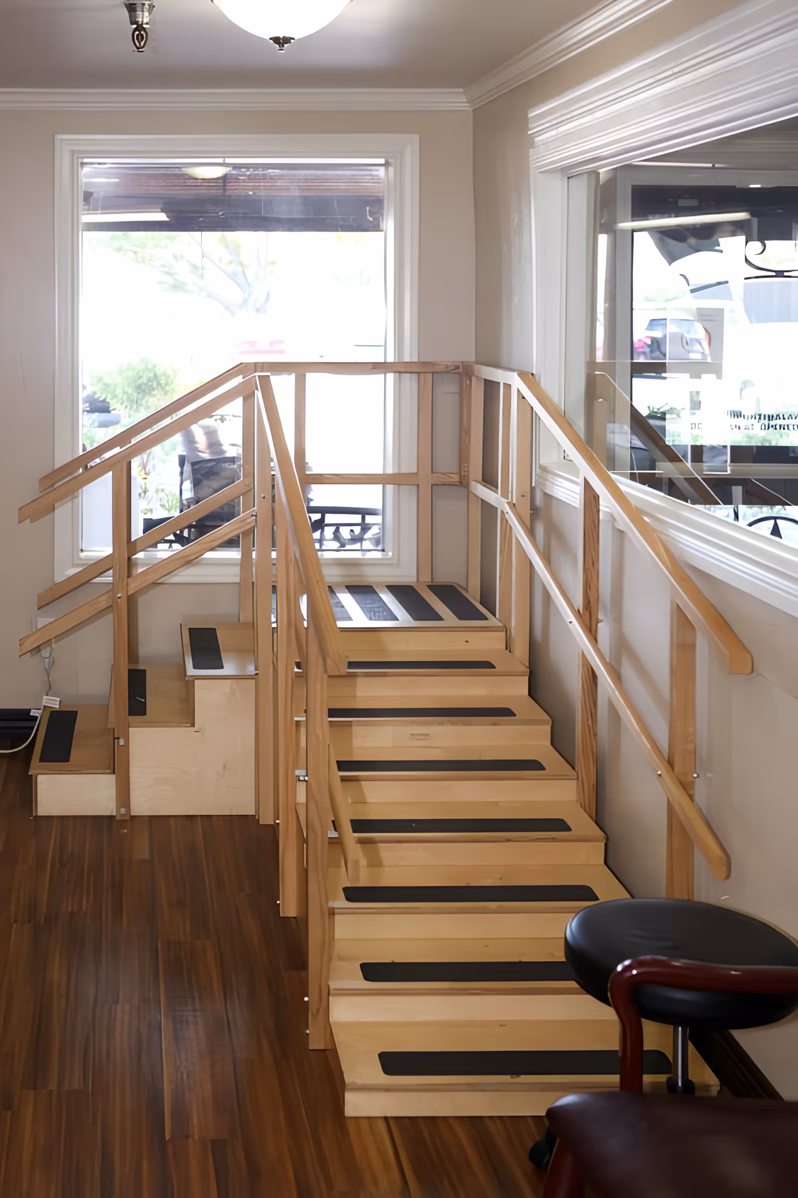 Interior view of a rehabilitation stair-training area with wooden practice stairs, handrails, and large windows.