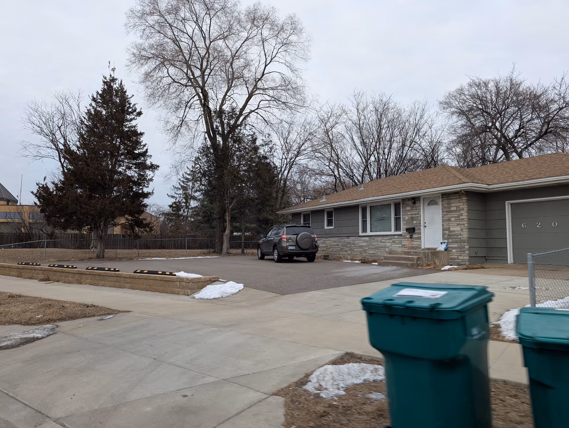 Single-story ranch-style house with a driveway, a parked SUV, and green trash bins in the foreground