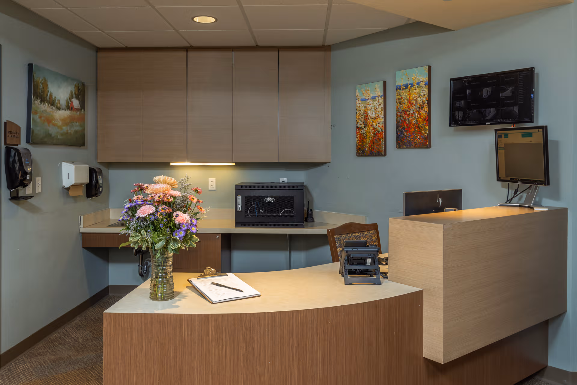 Reception desk with flowers, cabinets, and monitors in a clinic-like lobby area.