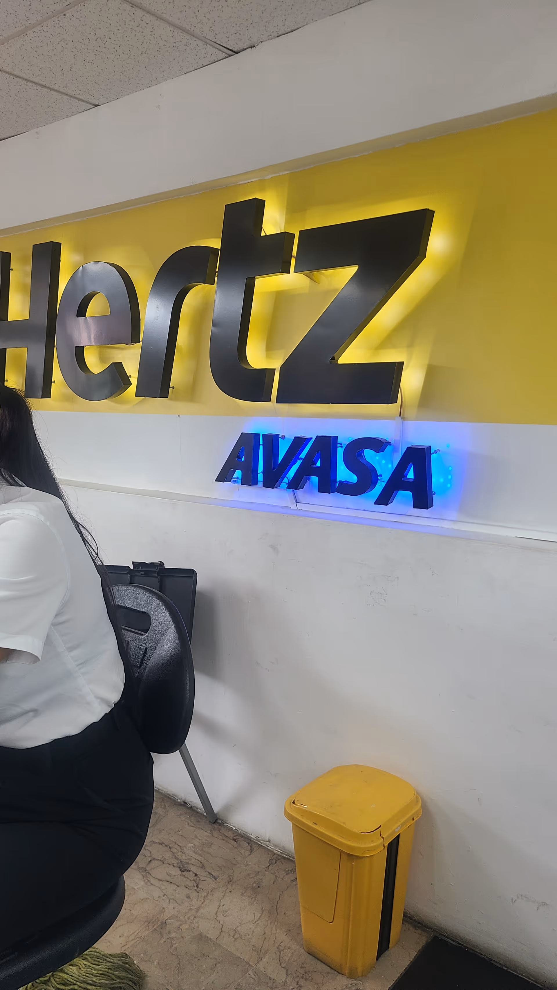 Interior lobby with yellow wall, illuminated Hertz Avasa signs, and a yellow trash bin.
