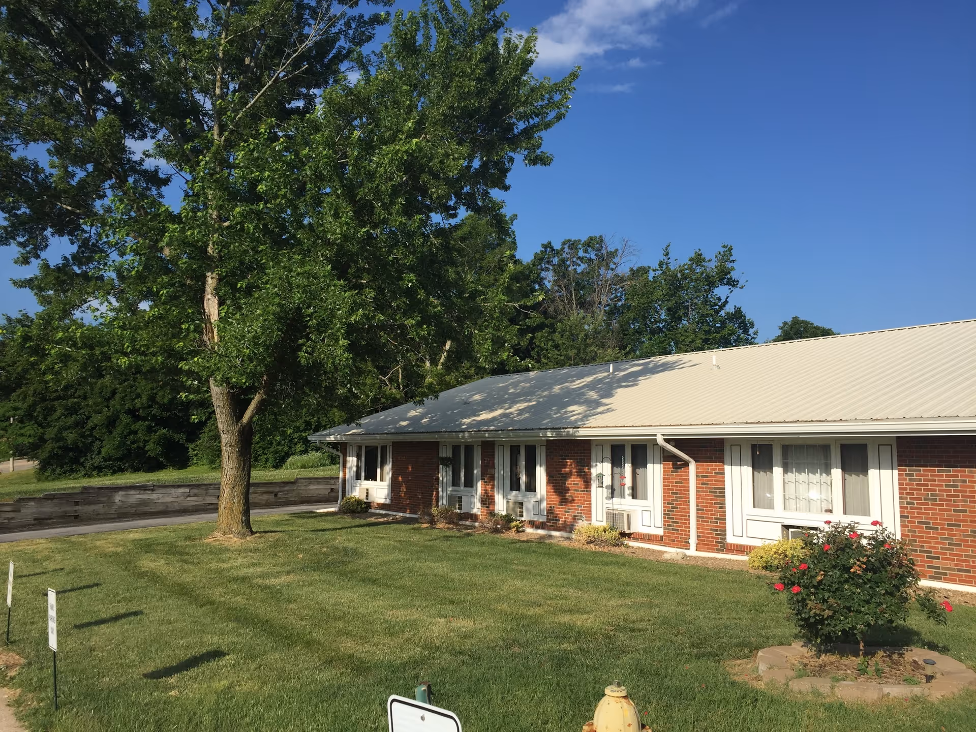 Single-story brick building with a light-colored metal roof, several windows, and doors facing a well-maintained grassy lawn with a tree and a flower bush. The sky is clear and blue.