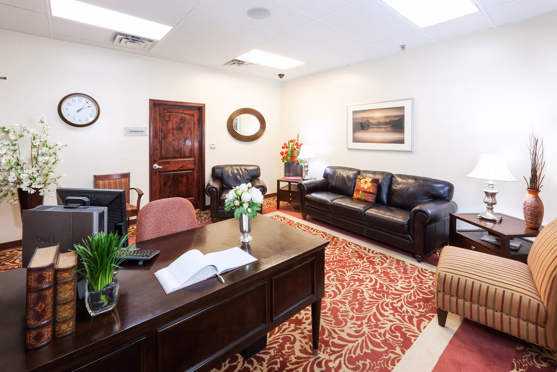 A well-lit office or reception area with a dark wooden desk in the foreground, featuring a computer, books, a plant, and an open book. Behind the desk is a seating area with a black leather sofa, a matching armchair, a striped armchair, side tables with lamps, a wall clock, a round mirror, and a framed landscape picture on the wall. The room has a patterned red and beige carpet and white walls.