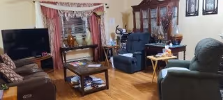 A cozy living room with wooden flooring featuring a brown leather sofa, a wooden coffee table with magazines underneath, two recliner chairs, a flat-screen TV on a stand, a wooden display cabinet with glass doors, and a window with pink curtains and a white lace valance.