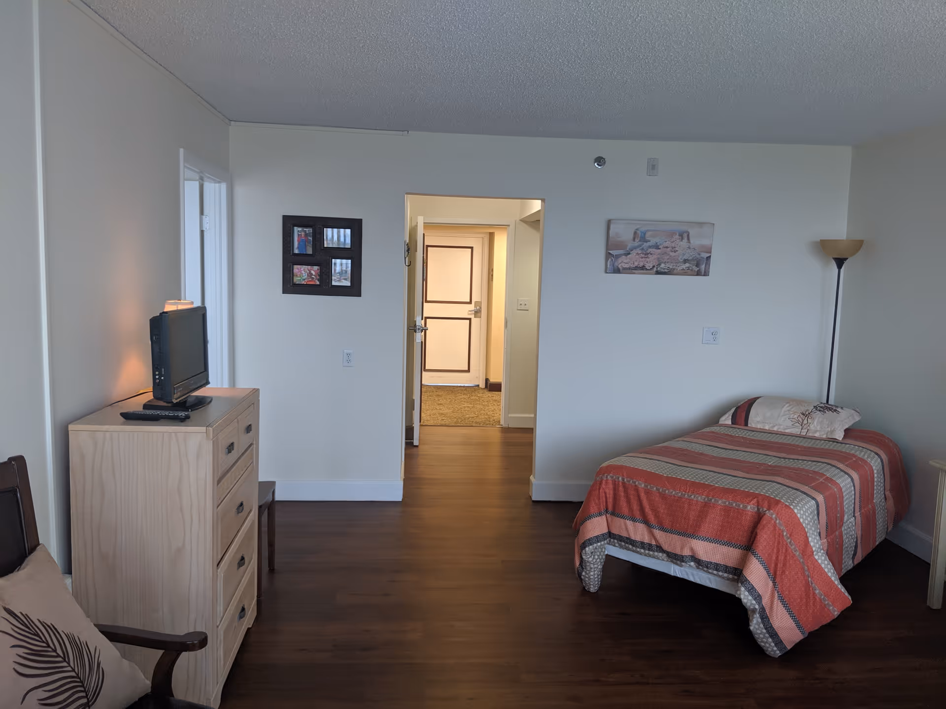 A furnished senior living bedroom with a single bed, dresser, TV, and hallway view.
