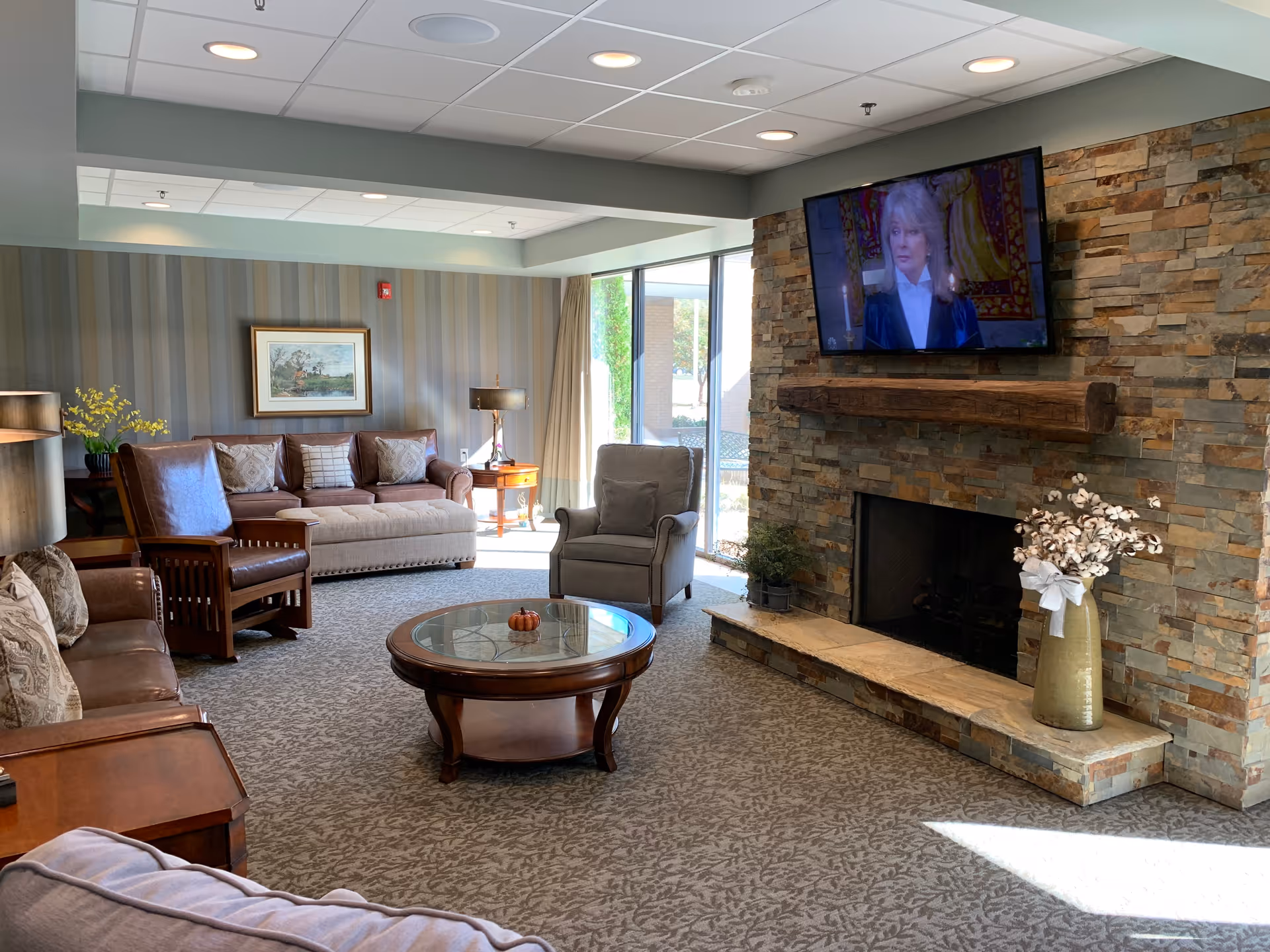 A cozy living room with a stone fireplace and a mounted flat-screen TV above it. The room features a beige sofa, a brown leather armchair, a gray armchair, a round glass coffee table with a small pumpkin decoration, and a side table with a lamp. There is a large window letting in natural light and a framed painting on the striped wallpapered wall.