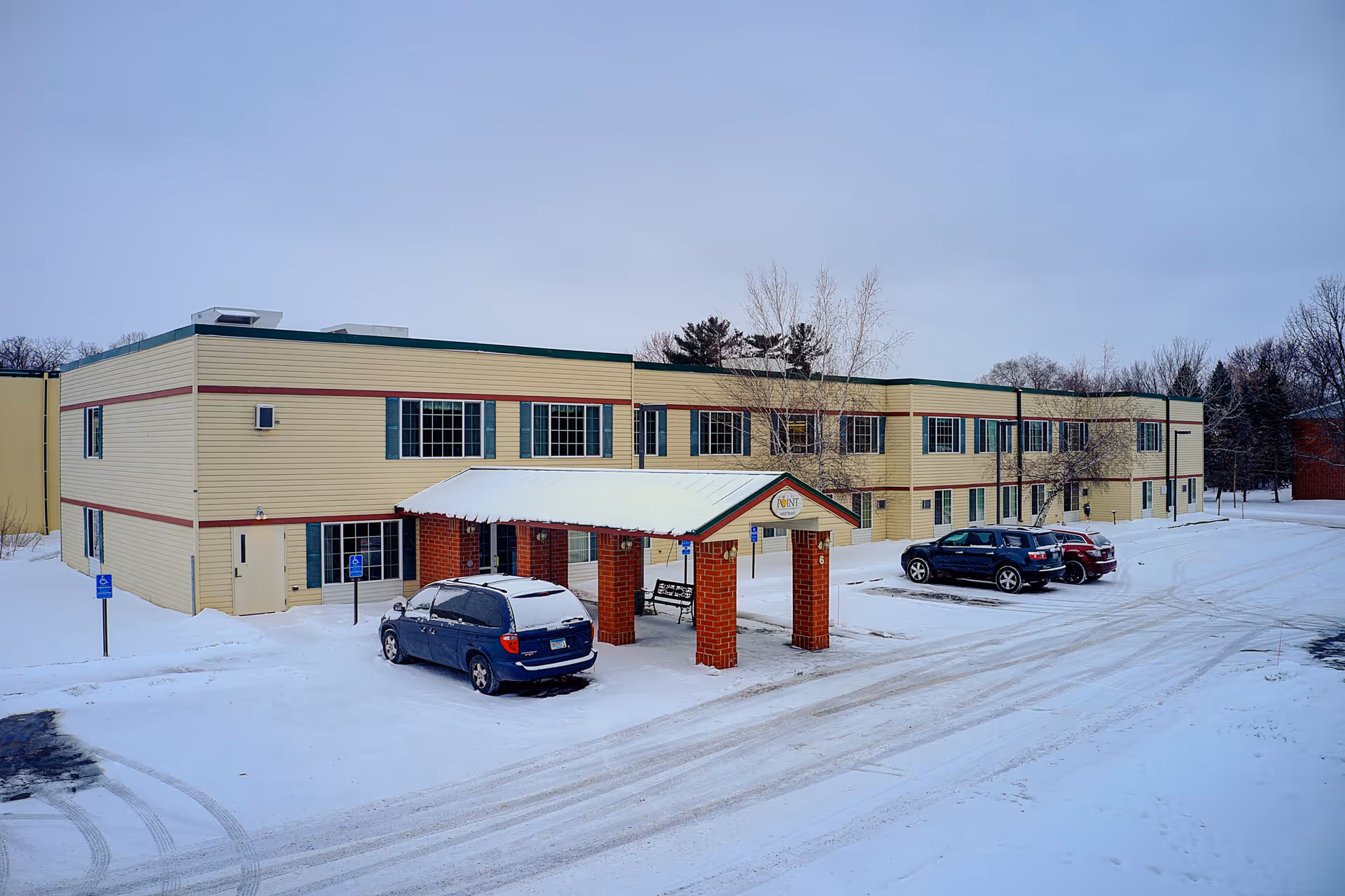 Exterior view of a two-story assisted living facility building named Nature's Point Assisted Living, surrounded by snow with a few parked cars and a covered entrance supported by brick pillars.
