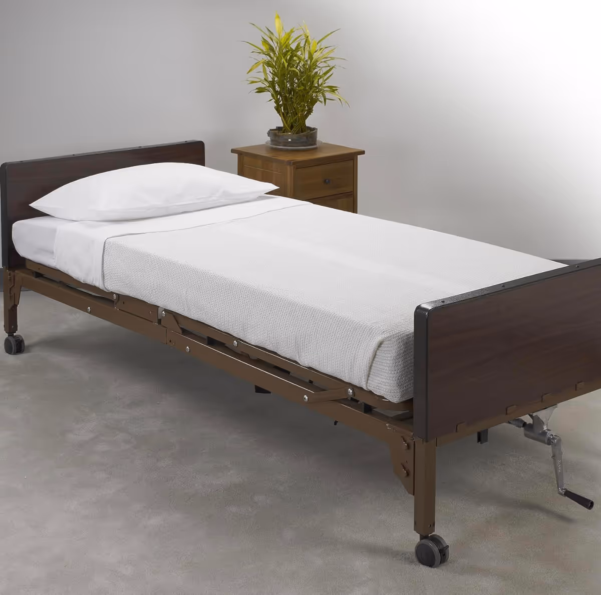 An adjustable hospital bed with white linens beside a wooden nightstand and plant.