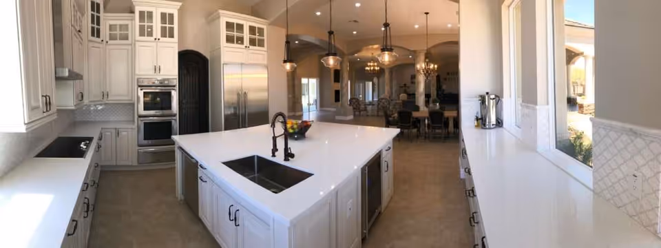 A spacious, modern kitchen with white cabinetry and countertops. The kitchen features a large central island with a built-in sink and faucet, stainless steel appliances including a double oven and refrigerator, and pendant lights hanging above the island. The kitchen opens into a dining and living area with chairs and a chandelier visible in the background. Large windows allow natural light to fill the space.