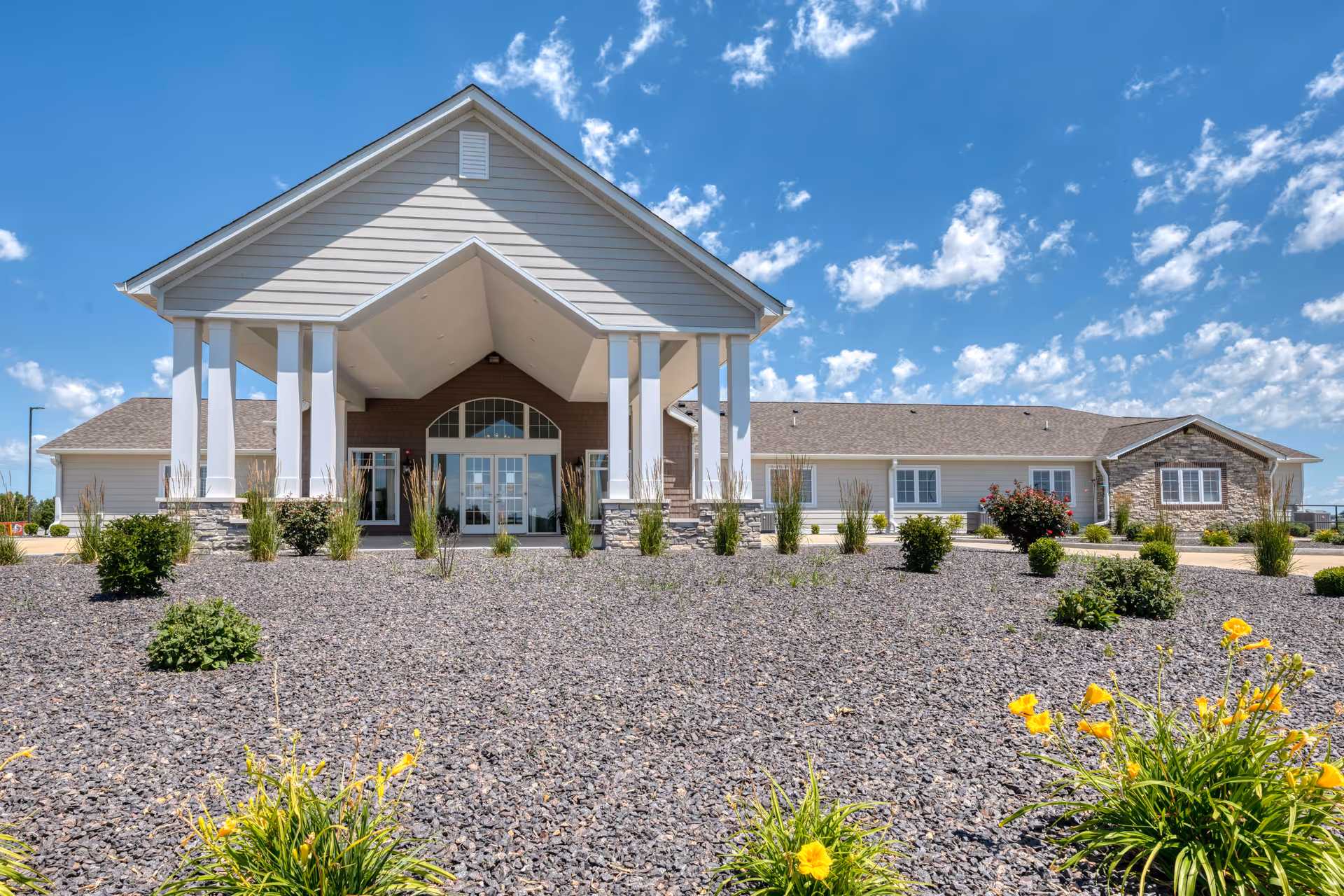 Front exterior view of Cedarhurst Senior Living of Bethalto building with a large covered entrance, stone and siding facade, landscaped garden with bushes and yellow flowers, under a blue sky with scattered clouds.