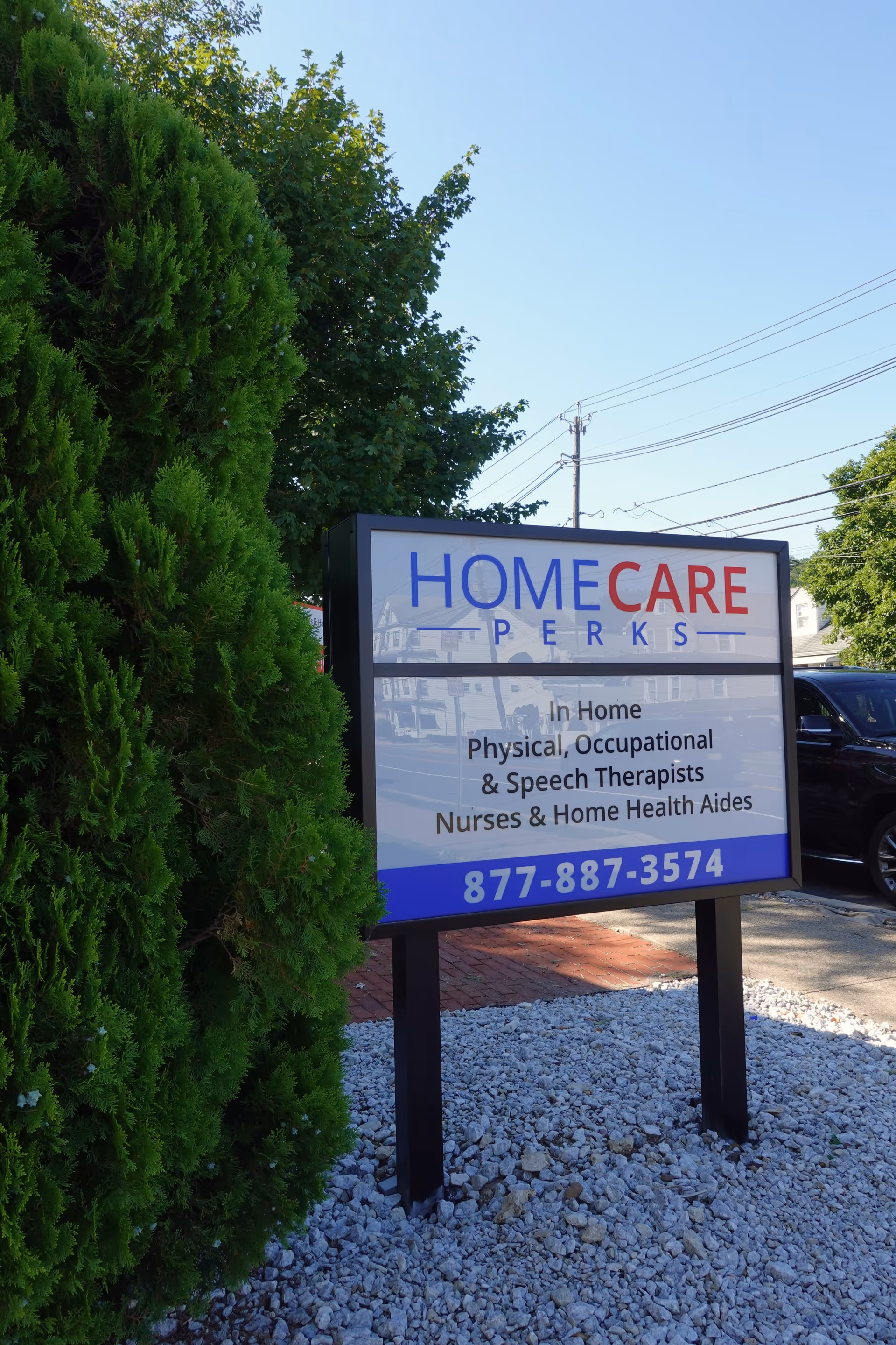 Outdoor sign for HomeCare Perks with services and phone number beside shrubs