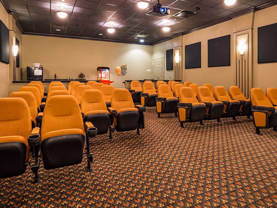 A small theater room with multiple rows of orange cushioned seats with black armrests and cup holders. The room has patterned carpet flooring, beige walls with black acoustic panels, and wall-mounted lights. At the back of the room, there is a popcorn machine and a small counter area with a microwave and sink. A projector is mounted on the ceiling.
