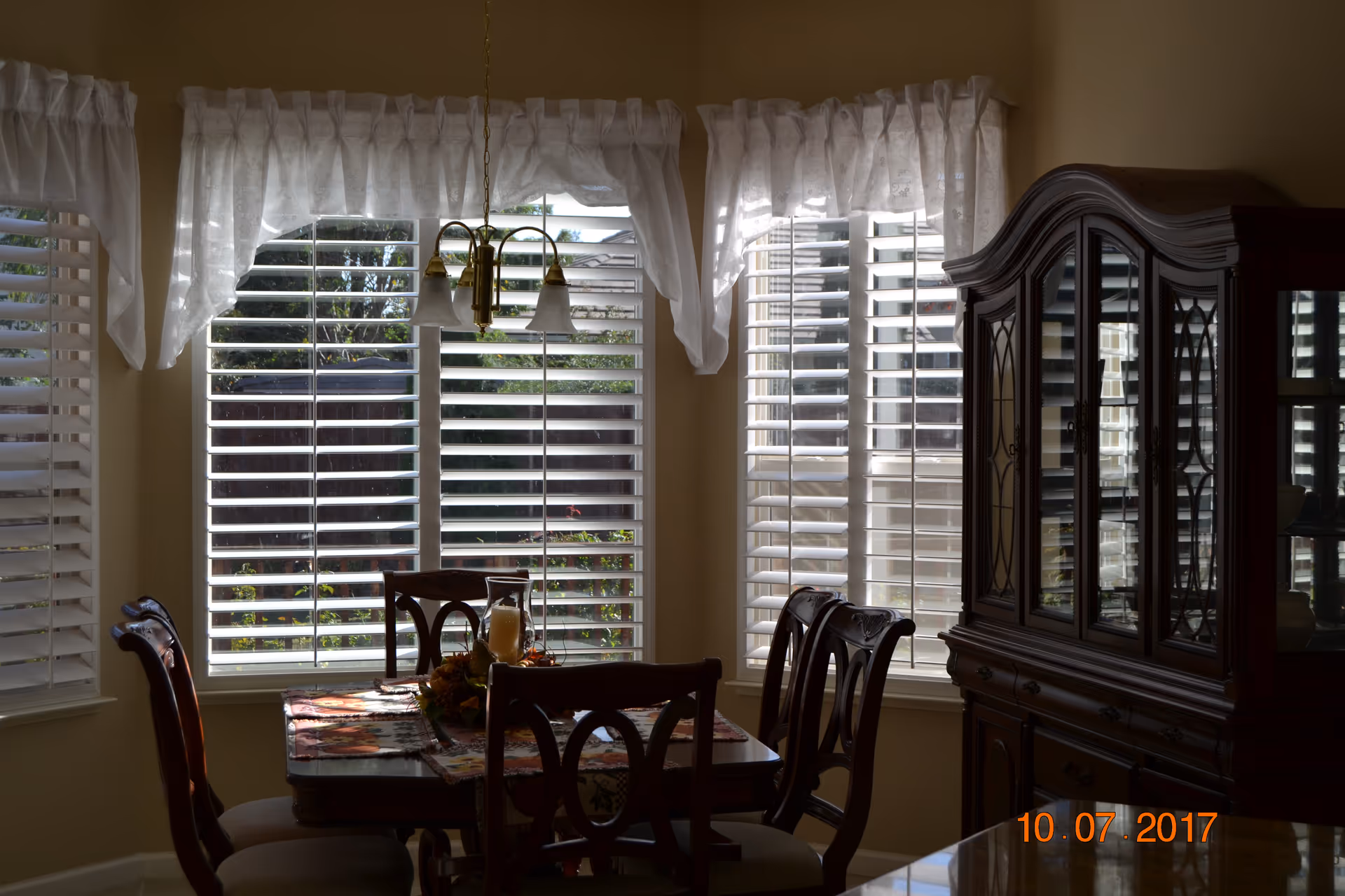 Sunlit dining room with wooden table, chairs, china cabinet, and white blinds.