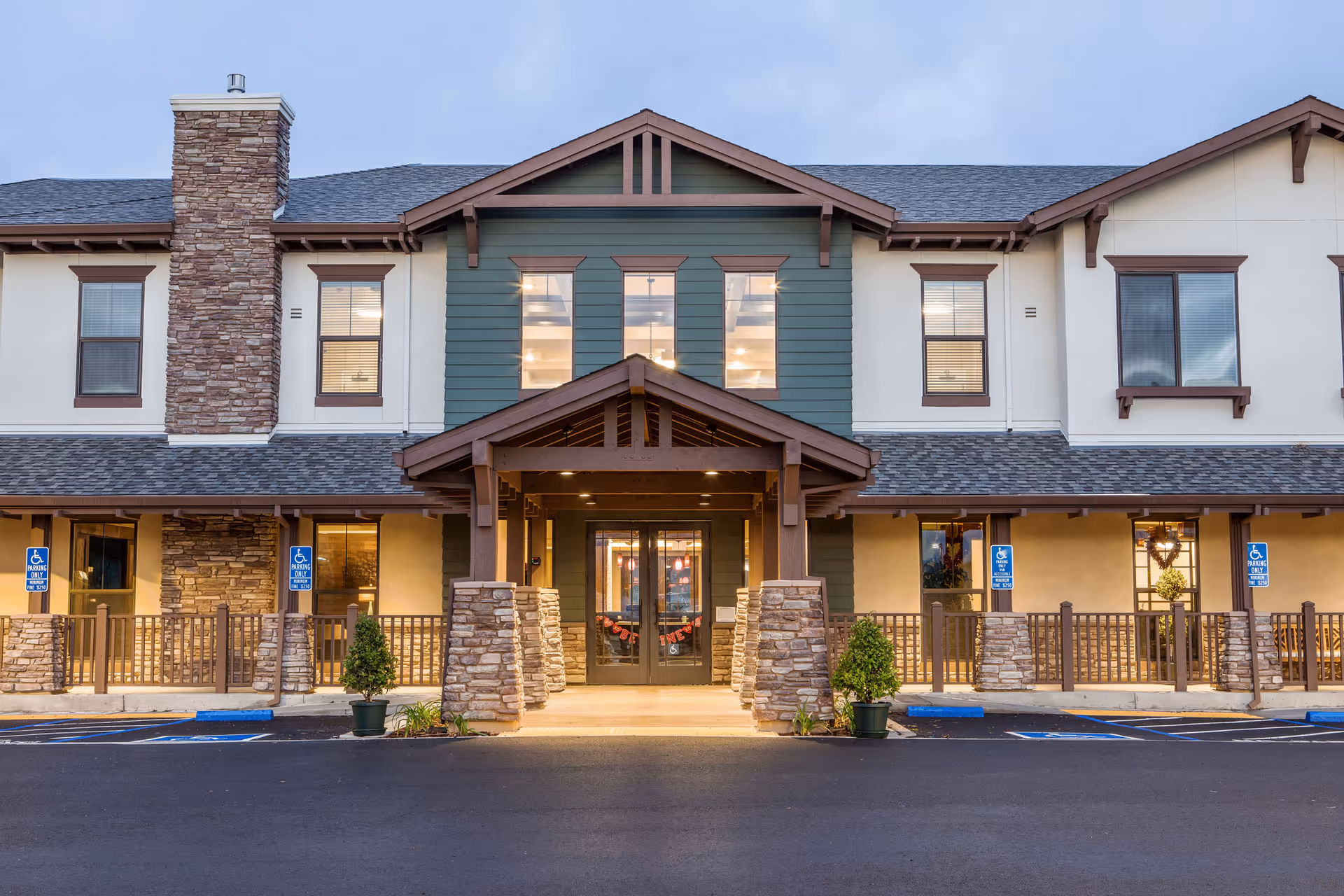 Front entrance of a two-story senior living building with a covered entry, stone columns, and accessible parking spaces.