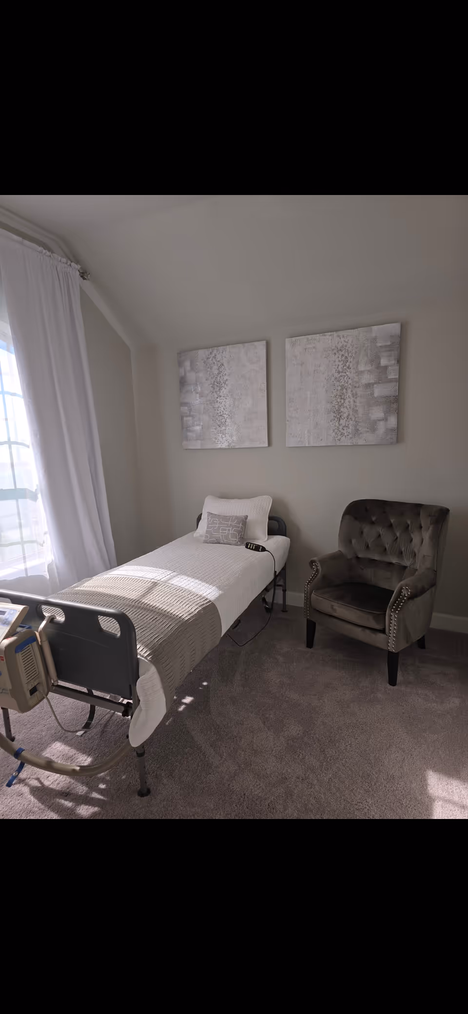 A hospital-style bed in a sunlit senior living bedroom with a chair and abstract wall art.