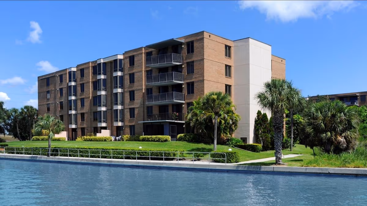 Four-story brick residential building with balconies facing a canal, landscaped grass and palm trees under a blue sky.