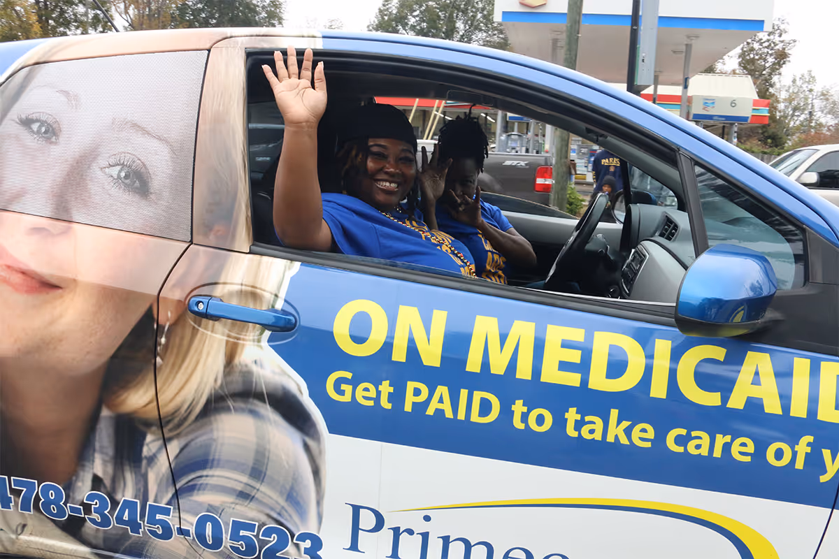 Two smiling women in a blue van with Primecare advertising and a large face graphic.