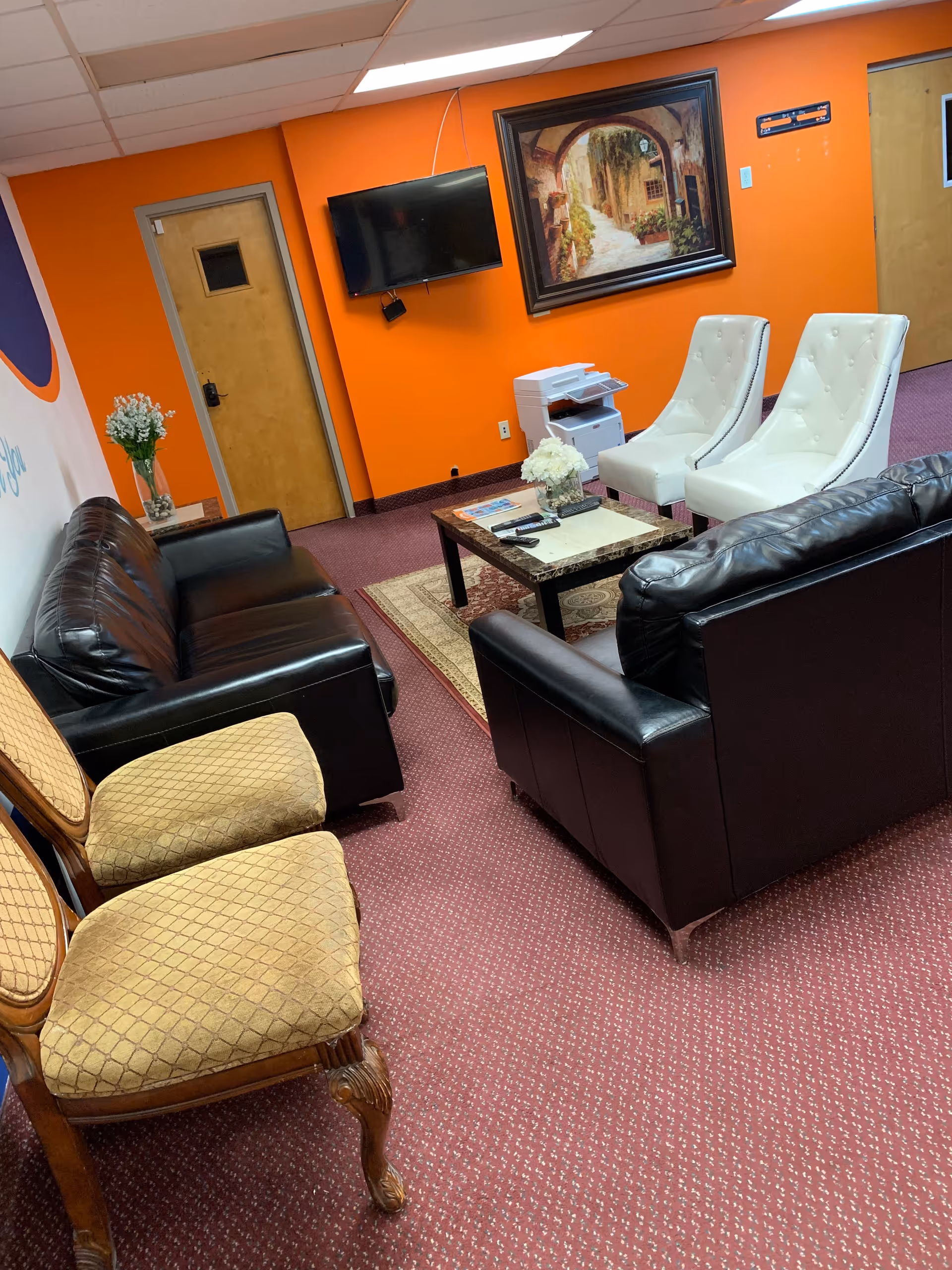 Common lounge area in a senior facility with orange walls, leather sofas, white chairs, and TV.