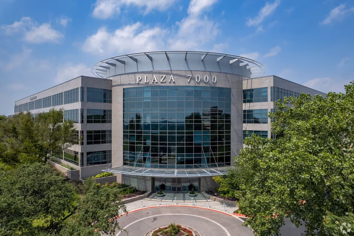 Exterior view of a modern building with a glass entrance labeled Plaza 7000, surrounded by trees.