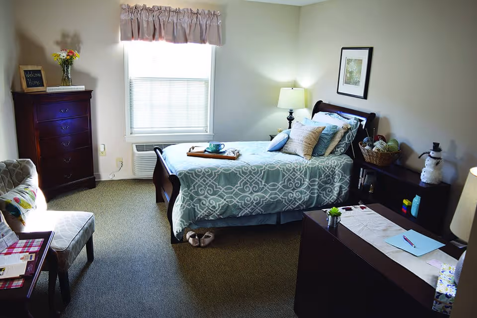 A cozy bedroom with a single bed covered in a light blue and white patterned bedspread. The bed has several pillows and a wooden tray with a cup and saucer on it. There is a wooden dresser with a vase of flowers and a small framed sign that says 'Welcome Home' near a window with blinds and a pink valance. A cushioned chair with a colorful pillow is positioned near the dresser. A nightstand with a lamp, a basket with yarn, and a small snowman decoration is next to the bed. A desk with a lamp, some papers, and a small plant is in the foreground.