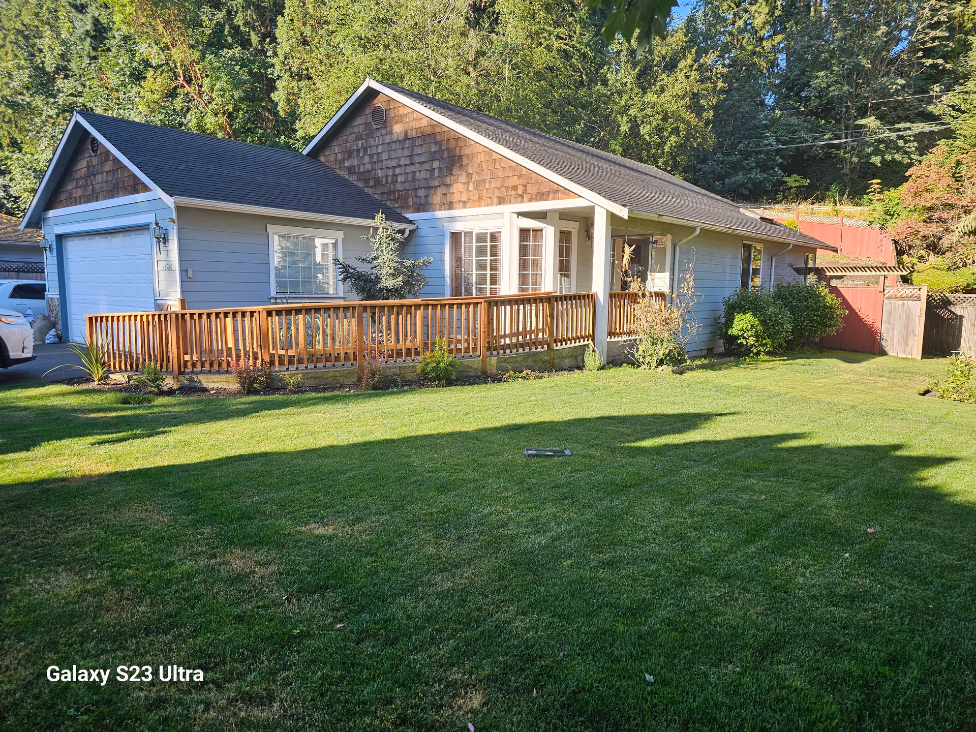 Blue single-story house with a wooden deck, green lawn, and surrounding trees