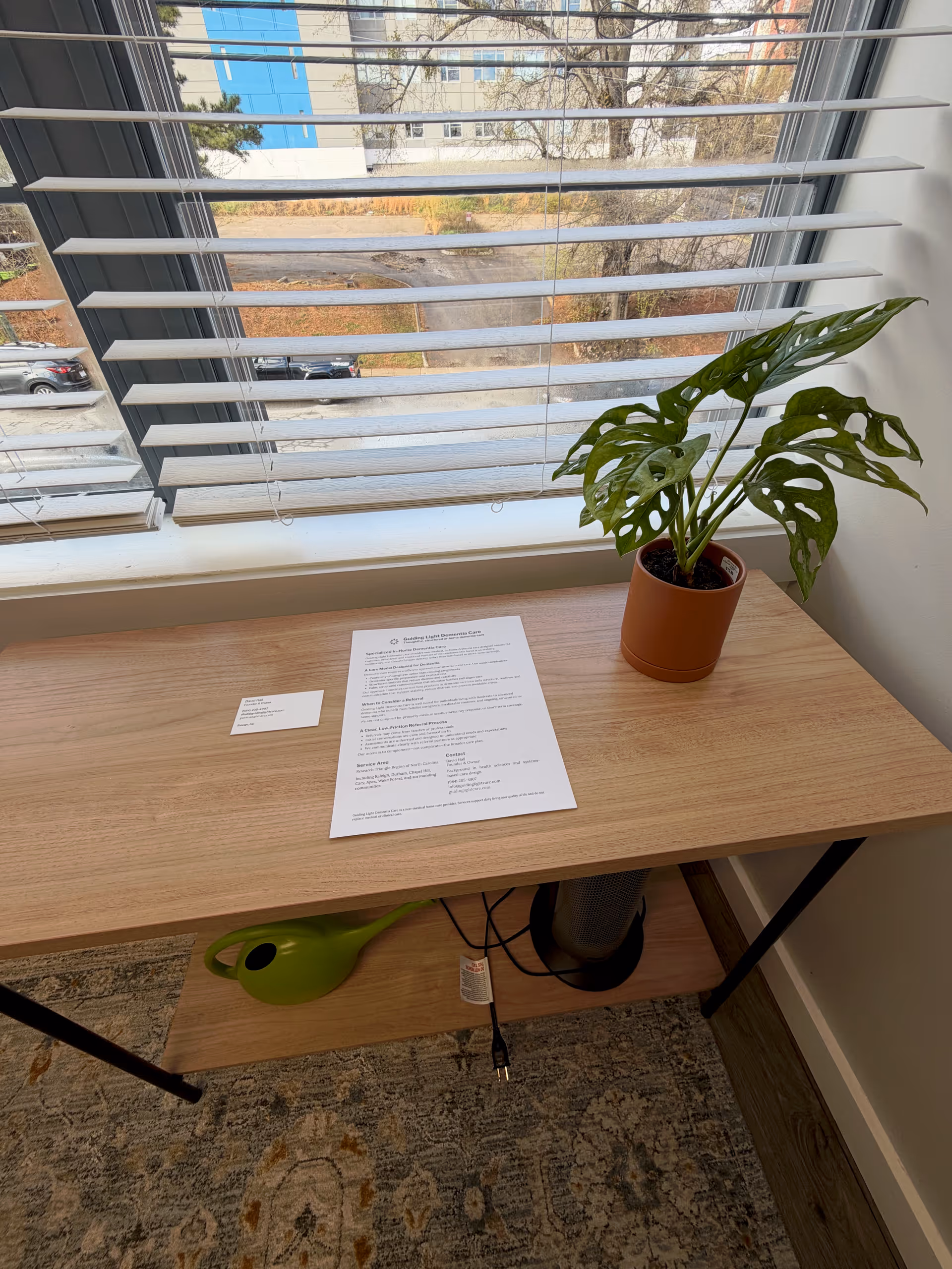 Sunlit desk near a window with blinds, a potted plant, and paperwork.