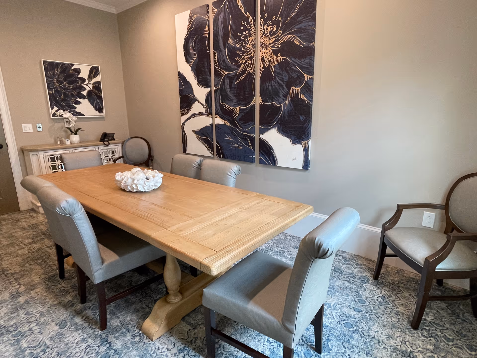 A dining room with a wooden rectangular table surrounded by six upholstered chairs. The room features a patterned carpet, a sideboard with a decorative plant and phone, and large floral artwork on the wall.