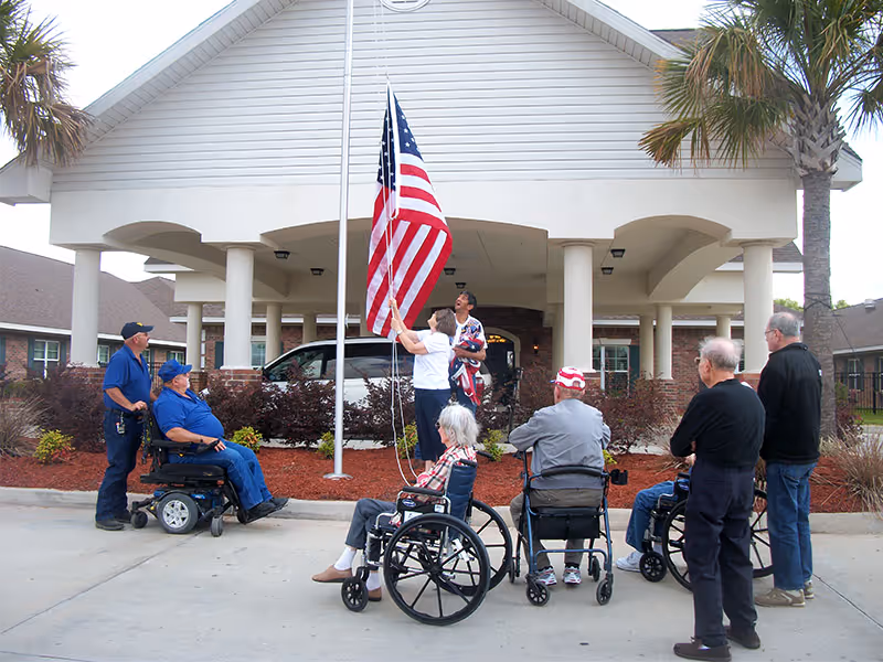 A group of elderly people, some in wheelchairs, gathered outside the entrance of a healthcare facility. Two individuals are raising an American flag on a flagpole in front of the building, which has a covered entrance with columns and palm trees nearby.