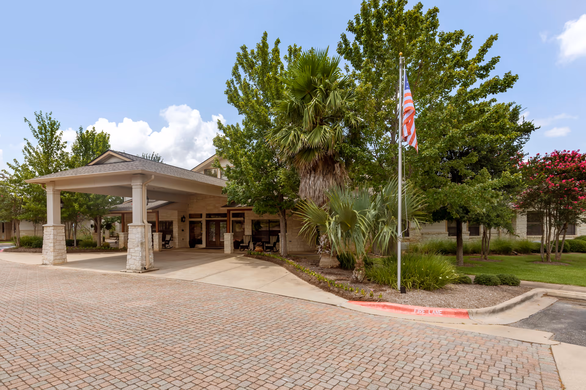 Front entrance of Brookdale Lohmans Crossing showing a covered porte-cochère, flagpole, palm trees, and a paved driveway.