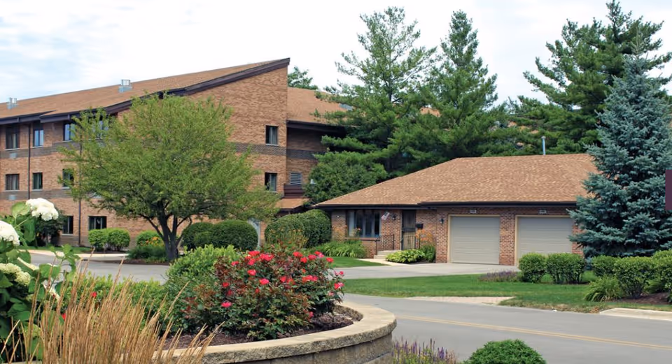 Siena on Brendenwood senior living building with brown roof, landscaped shrubs, trees, and a two-car garage.