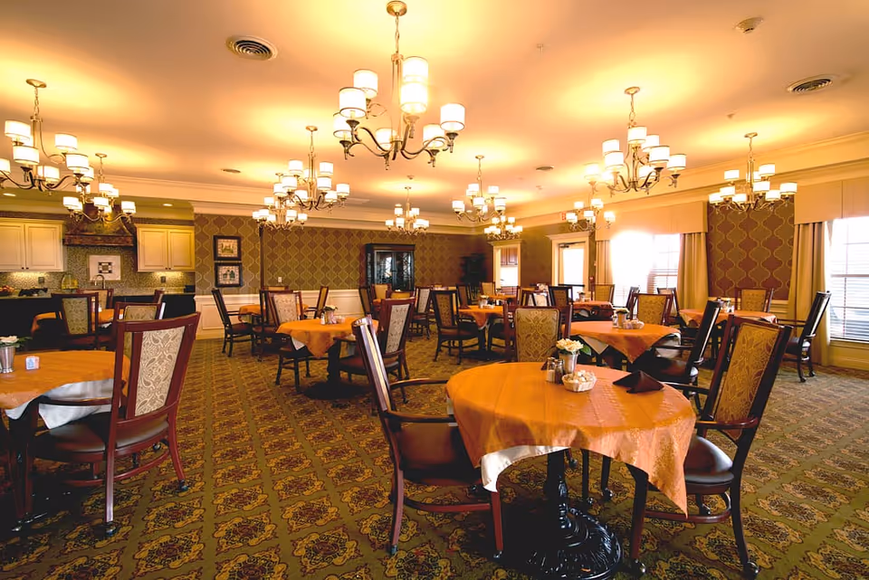 A spacious dining room with multiple round tables covered with orange tablecloths and surrounded by wooden chairs with patterned upholstery. The room features ornate chandeliers hanging from the ceiling, patterned carpet, and large windows with curtains allowing natural light to enter. The walls have decorative wallpaper and framed artwork, and there is a kitchen area visible in the background.