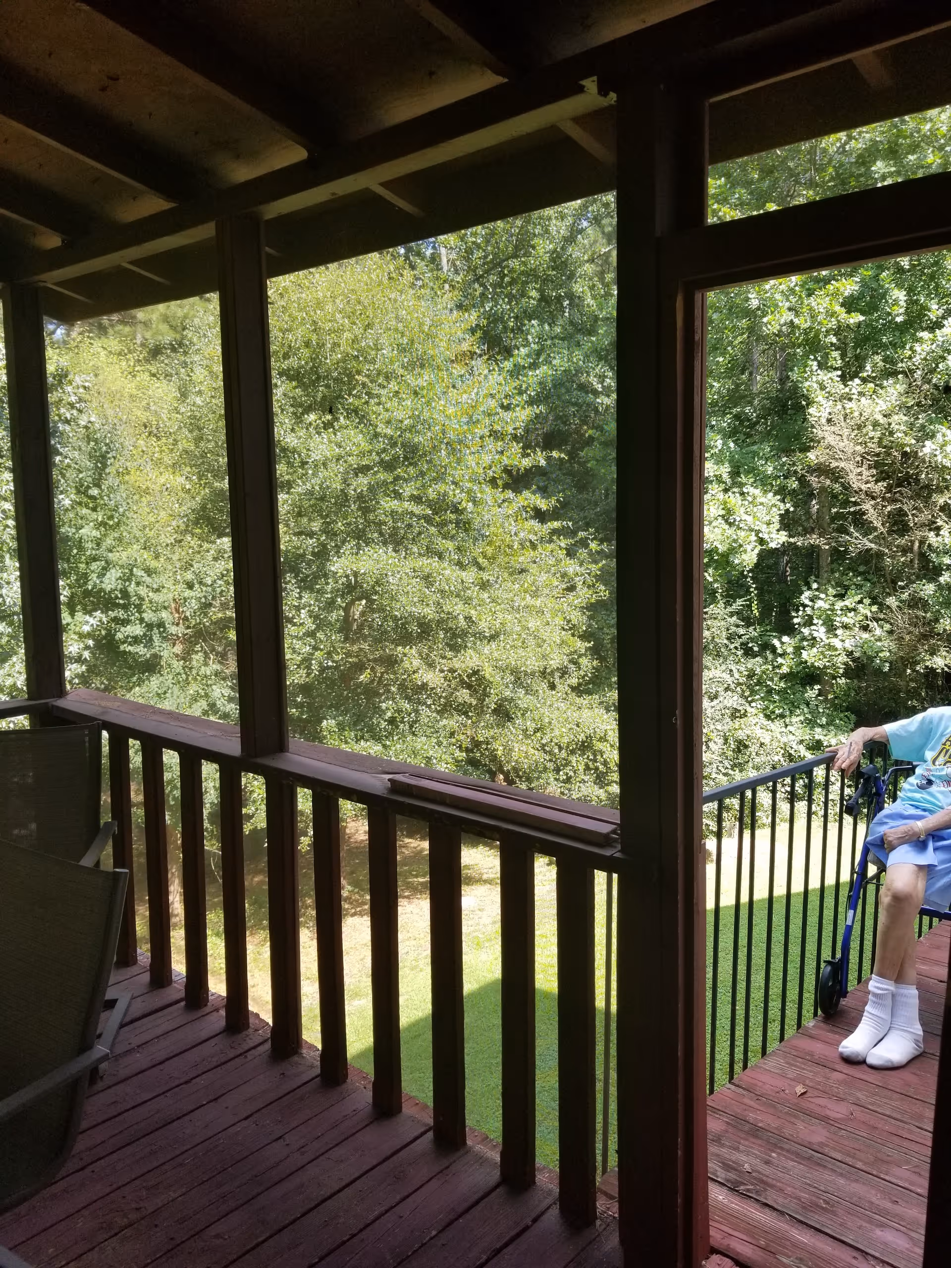 A wooden screened porch overlooking a green treed yard with an elderly person sitting at the doorway next to a walker.