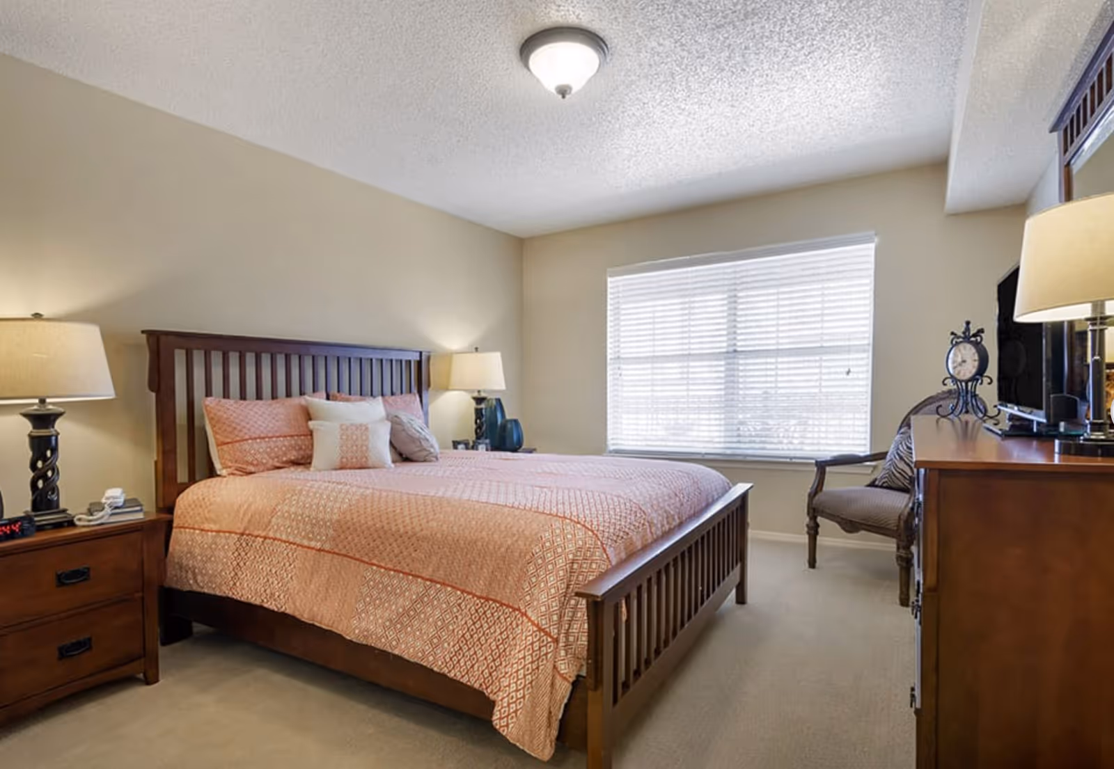 Well-lit bedroom with a wooden bed dressed in pink patterned bedding, bedside tables with lamps, a window with blinds, and a dresser with a chair.