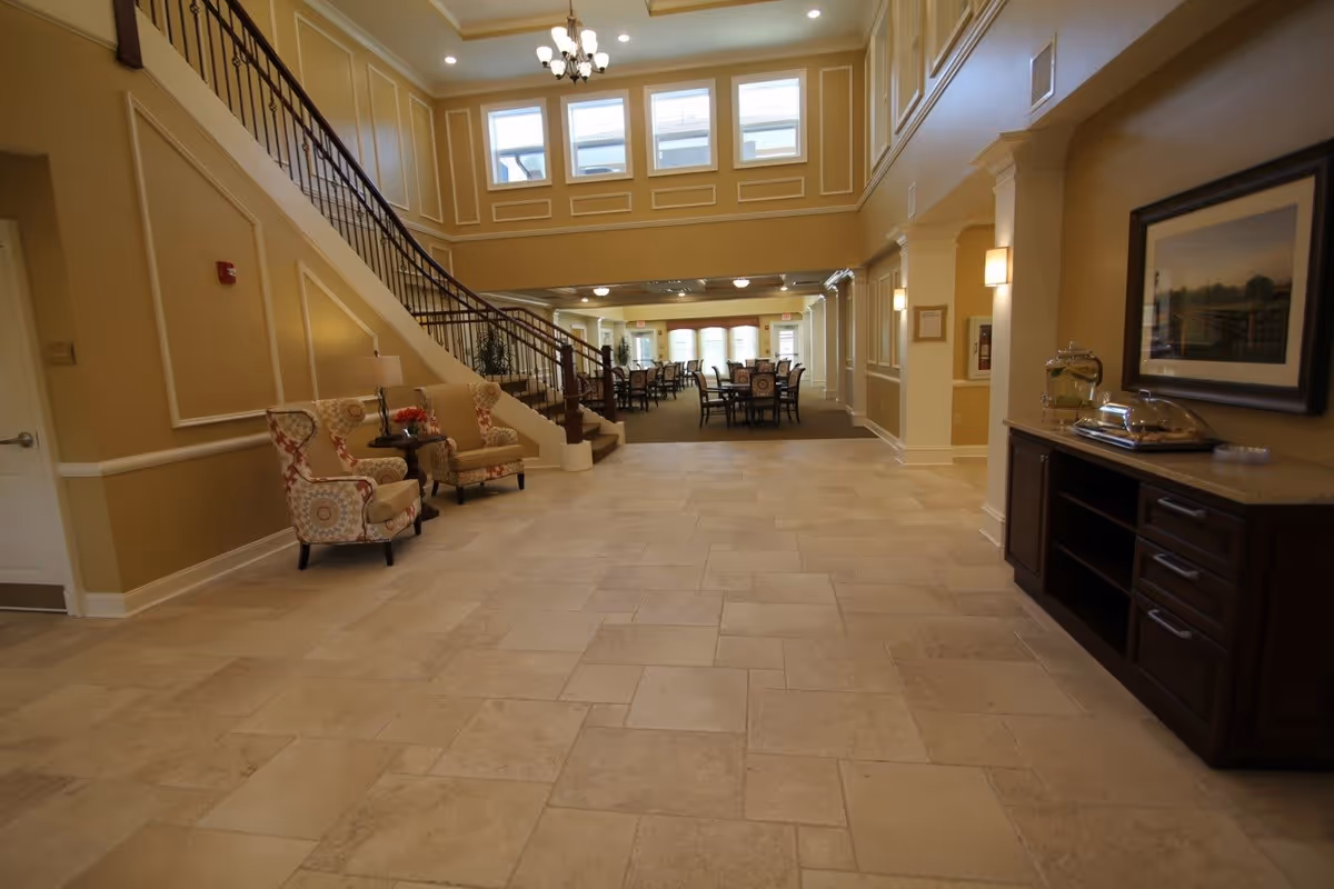 Spacious two-story lobby and common area with a tiled floor, curved staircase, seating and dining tables in the background.