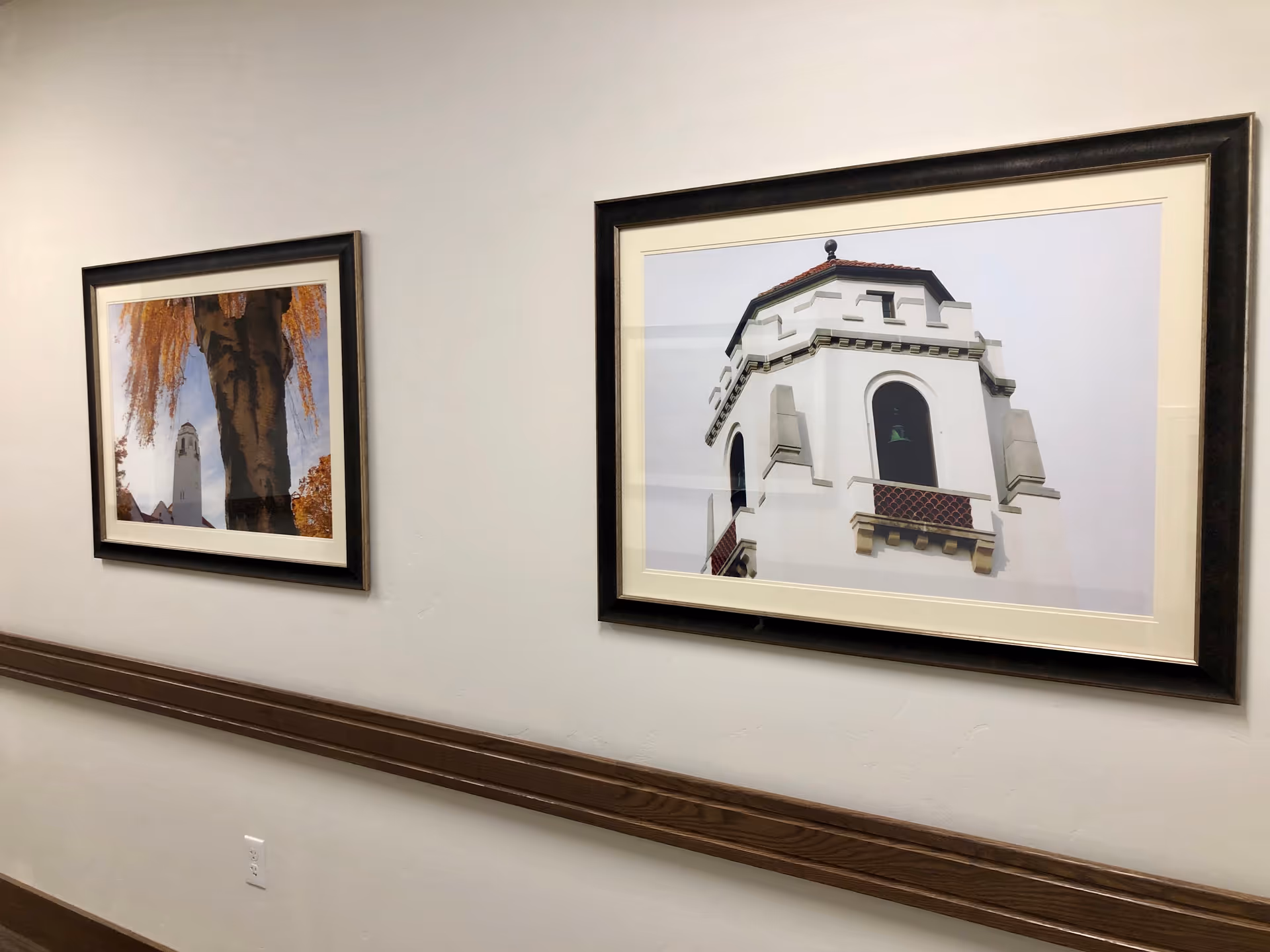 A hallway wall featuring two framed architectural photos above a wooden baseboard trim.