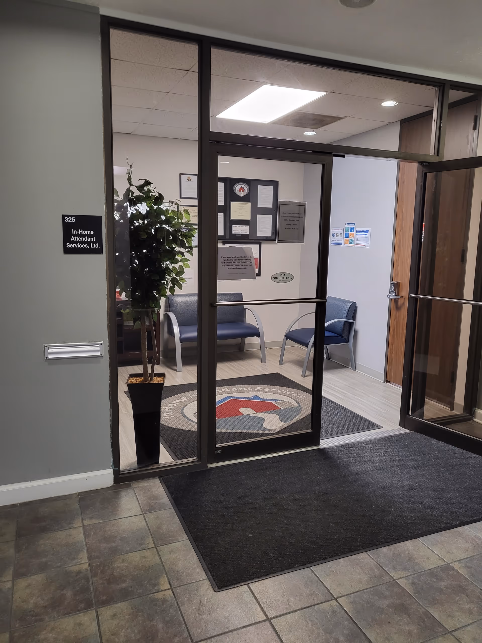 Interior entry lobby with glass doors, chairs, a plant, and a service logo rug.