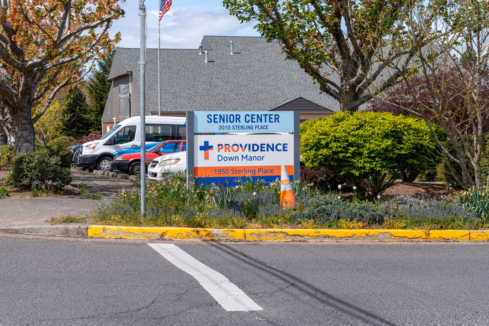 Sign for Providence Down Manor senior center at the entrance with parked vehicles, landscaping, and a building in the background.