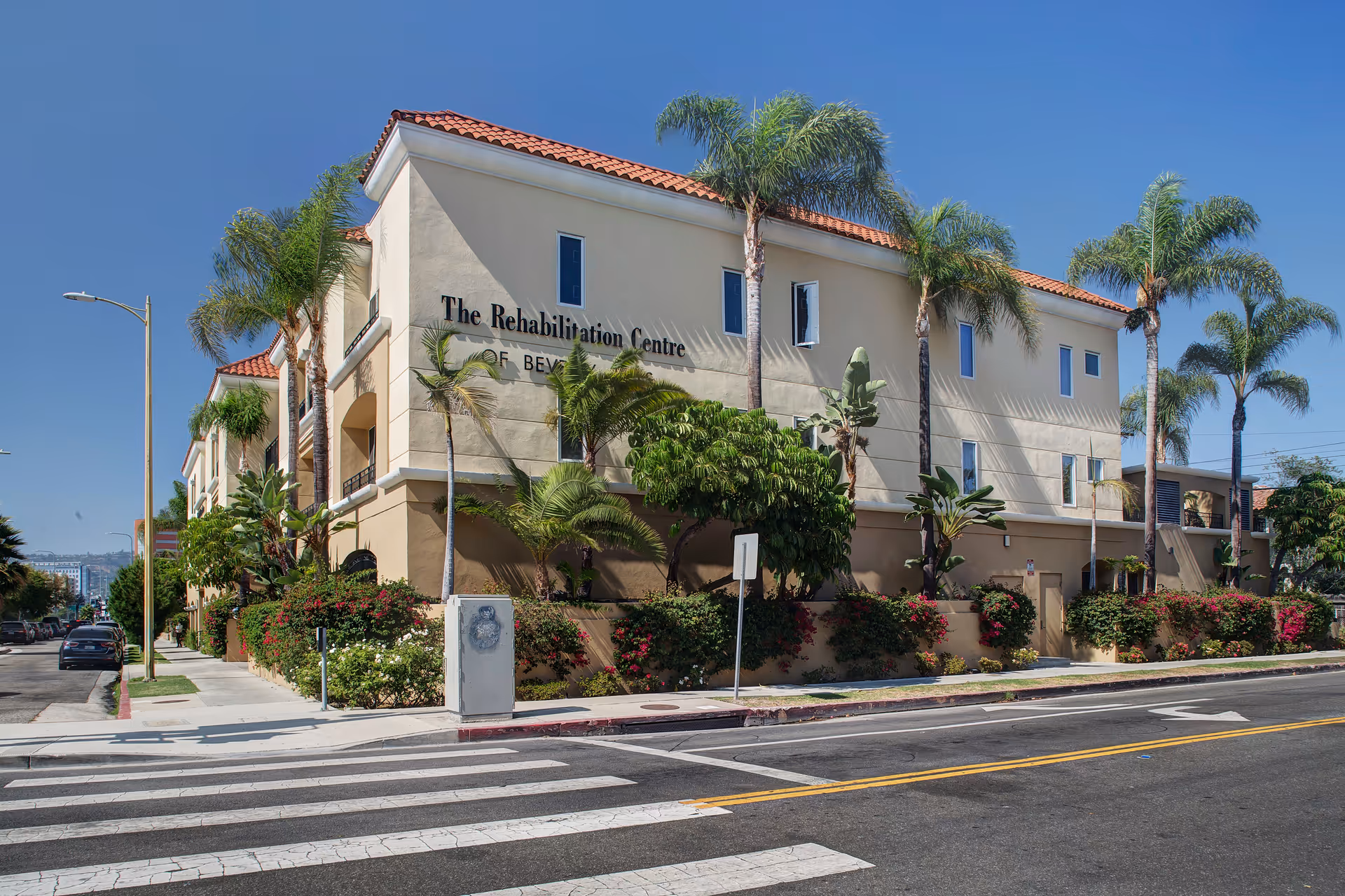 Two-story beige building with red-tiled roof and palm trees along the street.