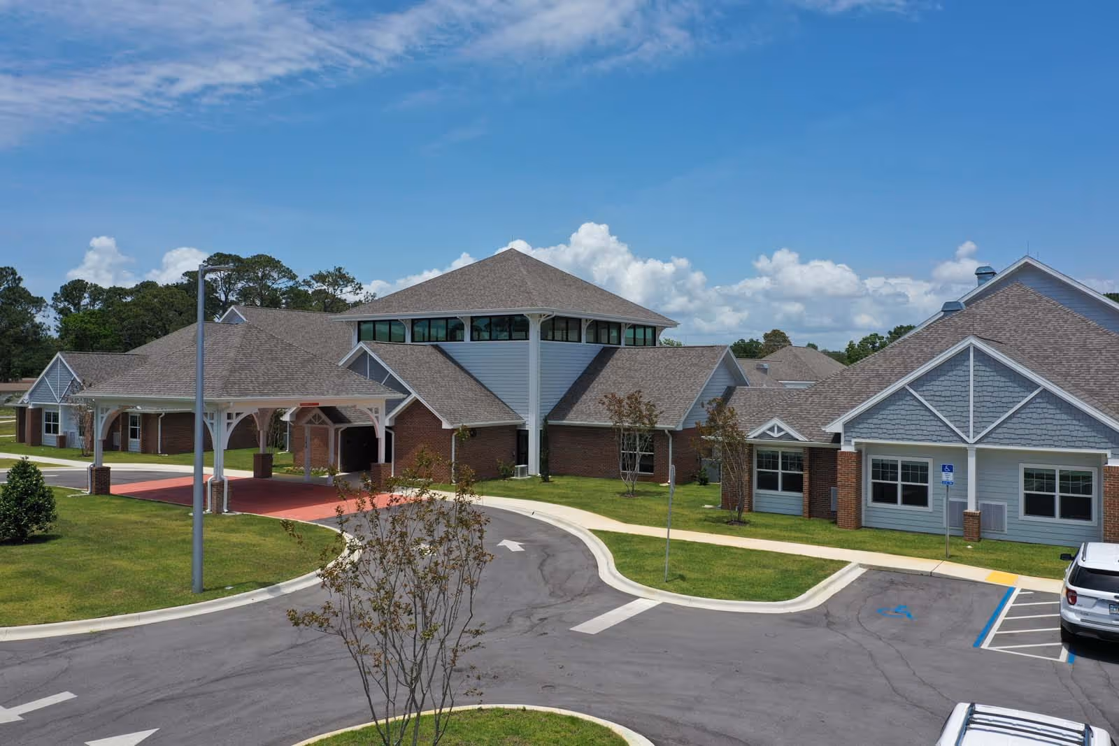 Front exterior of a single-story senior living facility with a covered entrance, drive loop, parking and landscaped lawns under a blue sky.