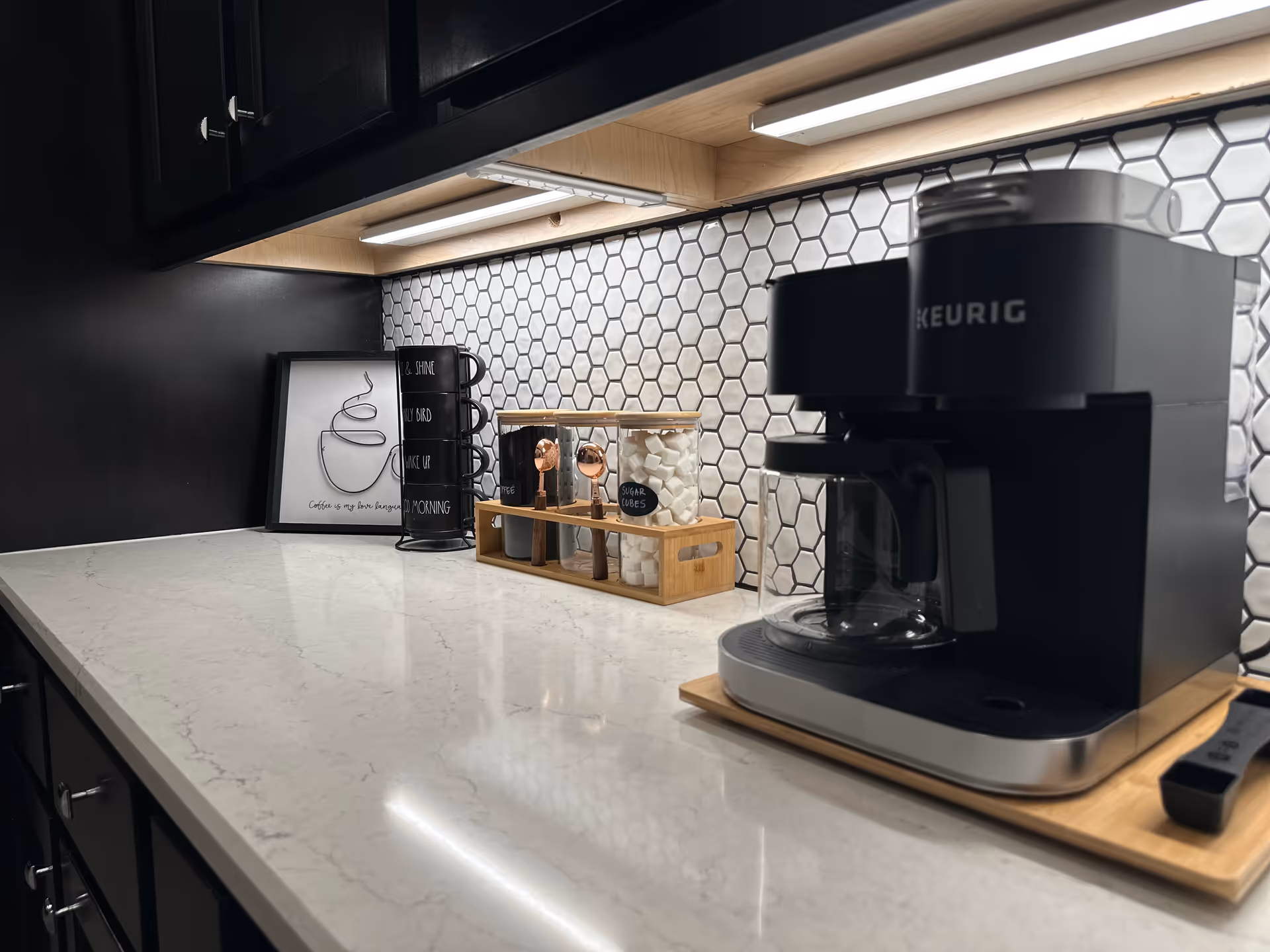 Modern kitchen counter with Keurig coffee maker, mug rack, framed art, and hex tile backsplash