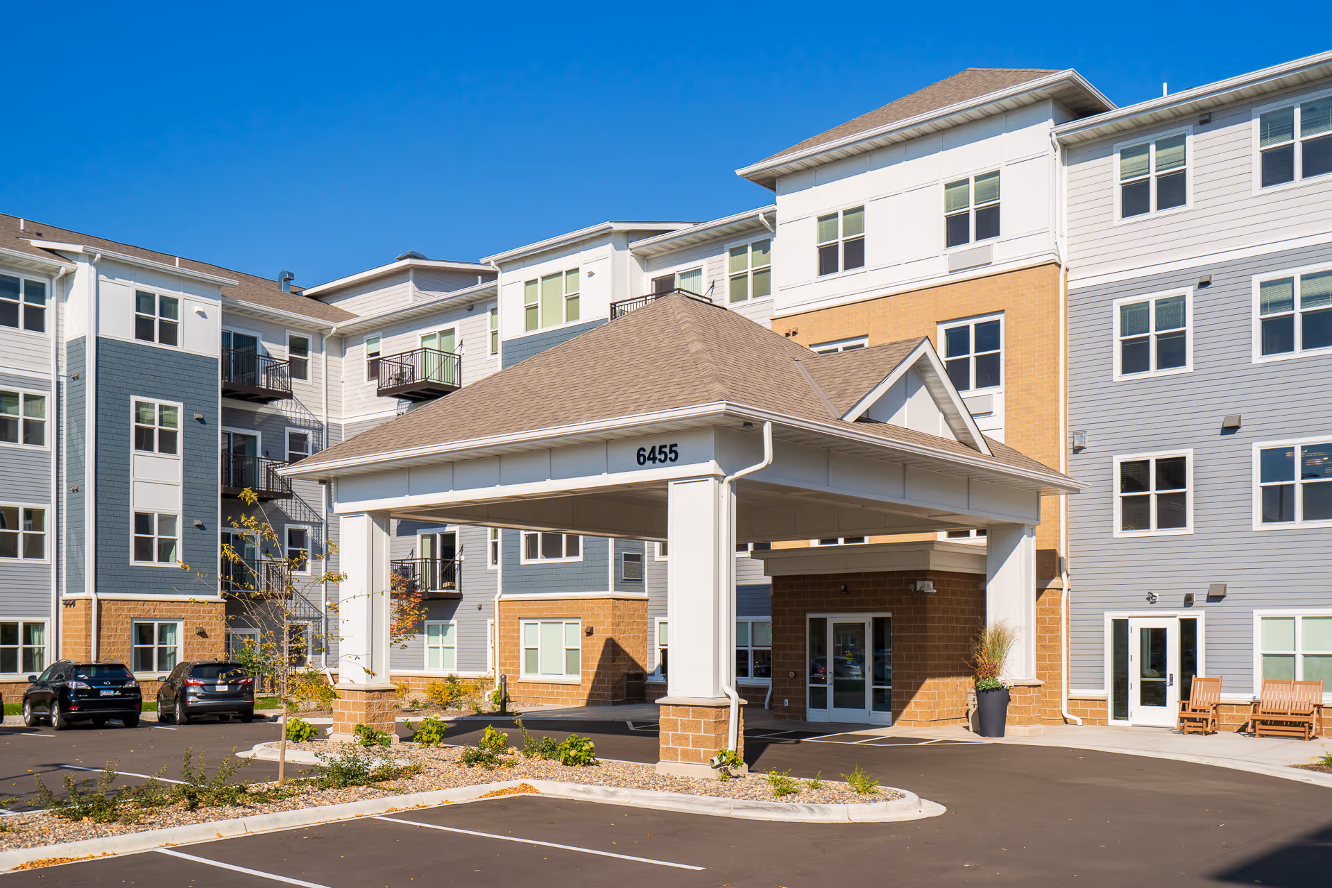 Exterior view of a modern multi-story senior living facility with a covered entrance and parking spaces in front. The building features a combination of light gray, white, and tan siding with multiple windows and balconies under a clear blue sky.