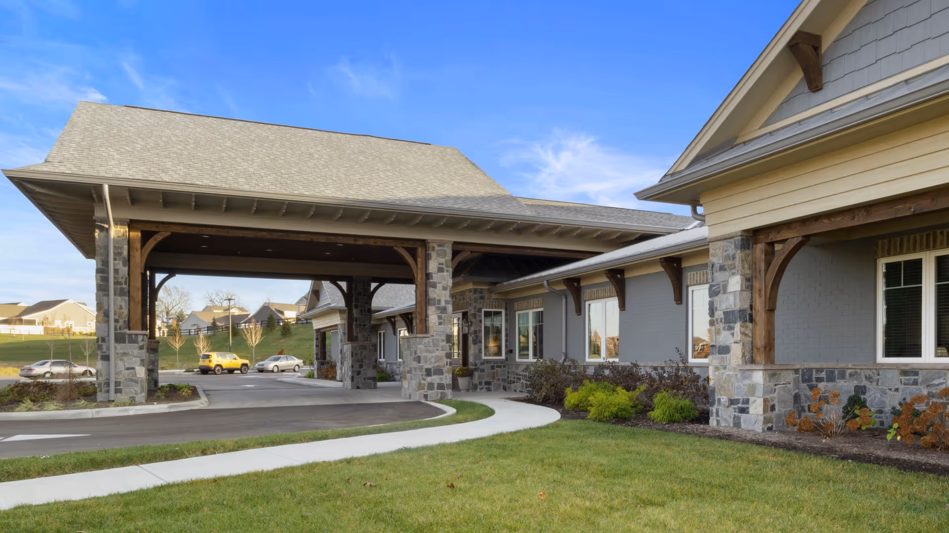 Exterior view of Beehive Homes of Liberty Township showing a covered entrance with stone pillars and wooden beams, a driveway, green lawn, and part of the building with windows and landscaping under a clear sky.