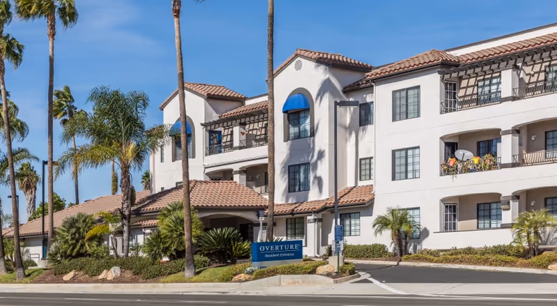 White multi-story Overture San Marcos senior living building with tiled roofs, palm trees, and blue sign.