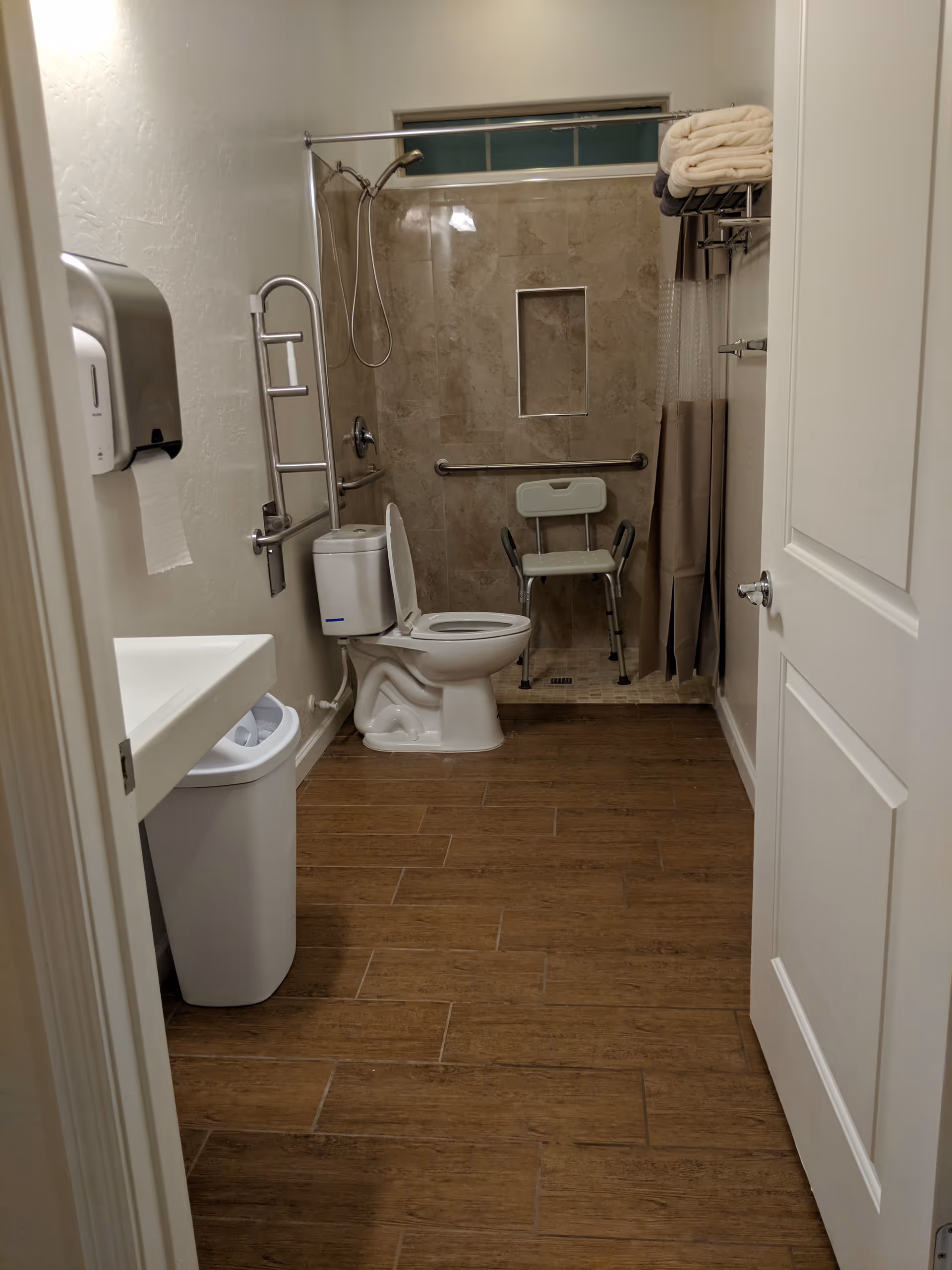 Accessible bathroom with toilet, grab bars, shower, sink, and a transfer chair.