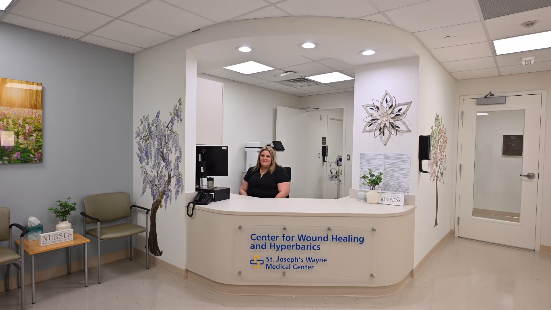 Reception desk in a medical center with staff member, chairs, and floral wall art.