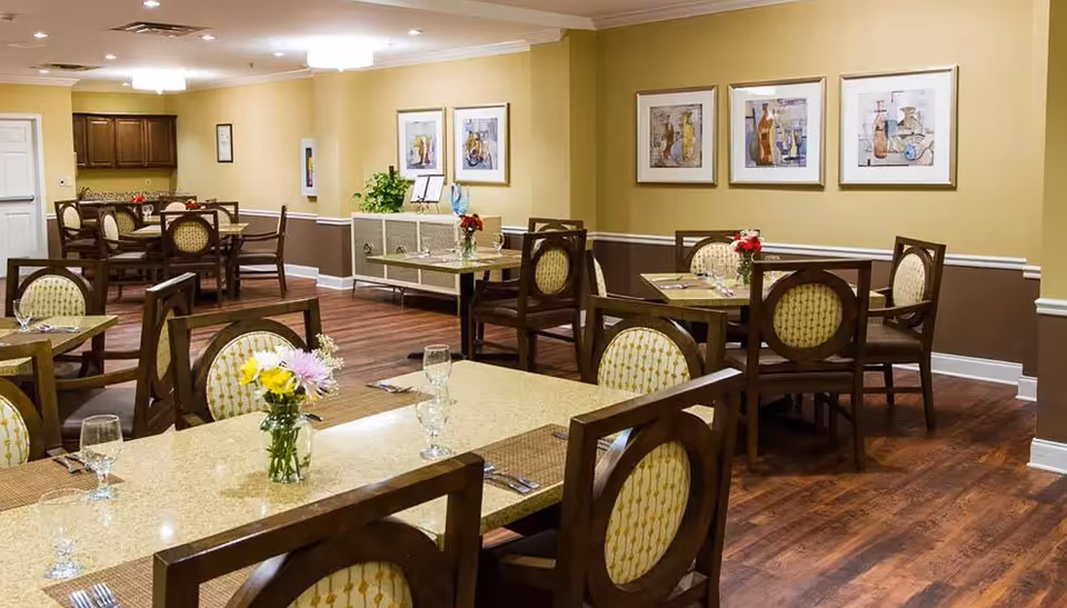 A dining room in a senior living facility with multiple tables and chairs arranged neatly. Each table has a small vase with flowers and place settings including glasses and utensils. The room has wooden flooring, beige and brown walls with framed artwork, and a sideboard with decorative items.
