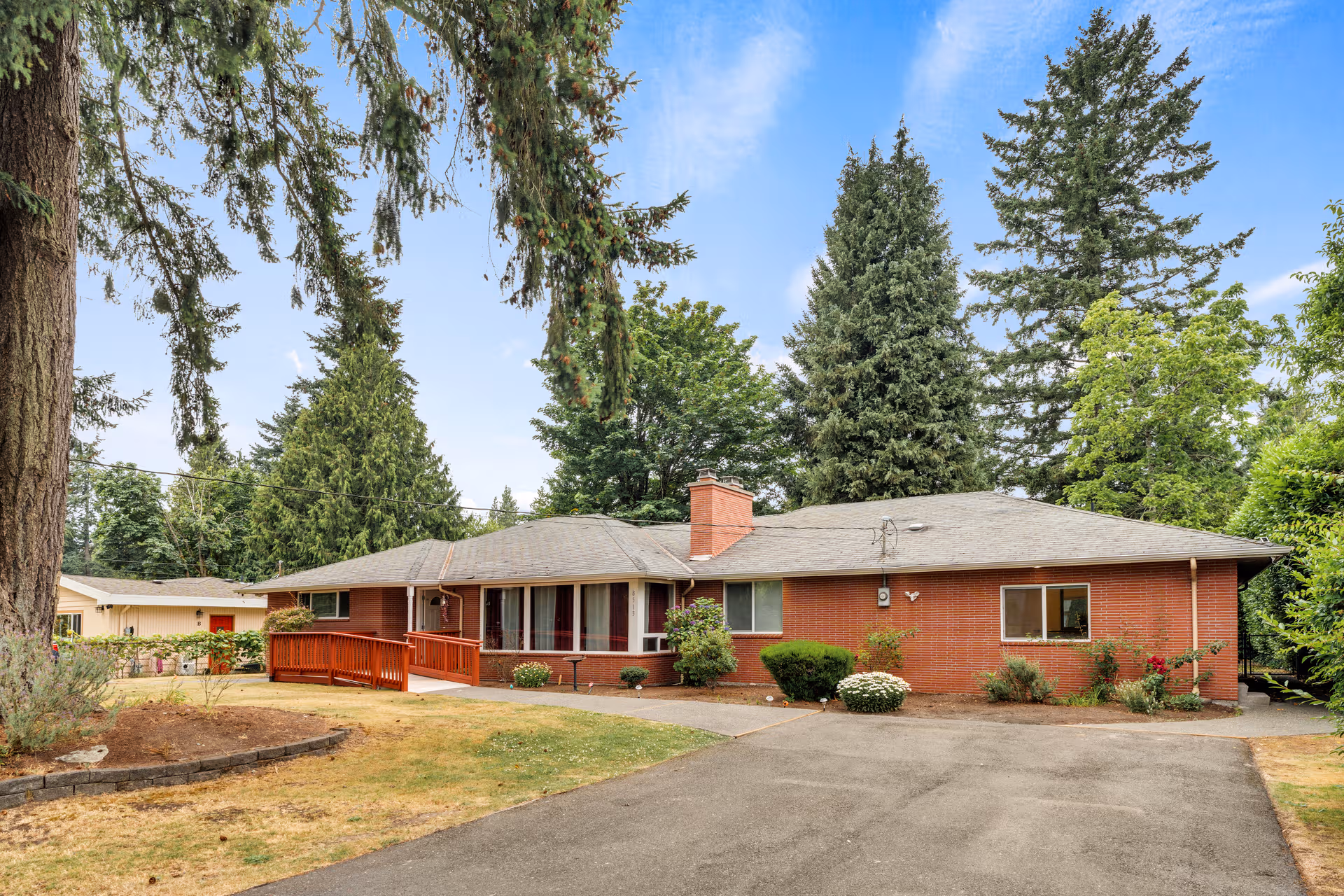 Single-story red brick senior living facility with a driveway, shrubs, and tall trees.
