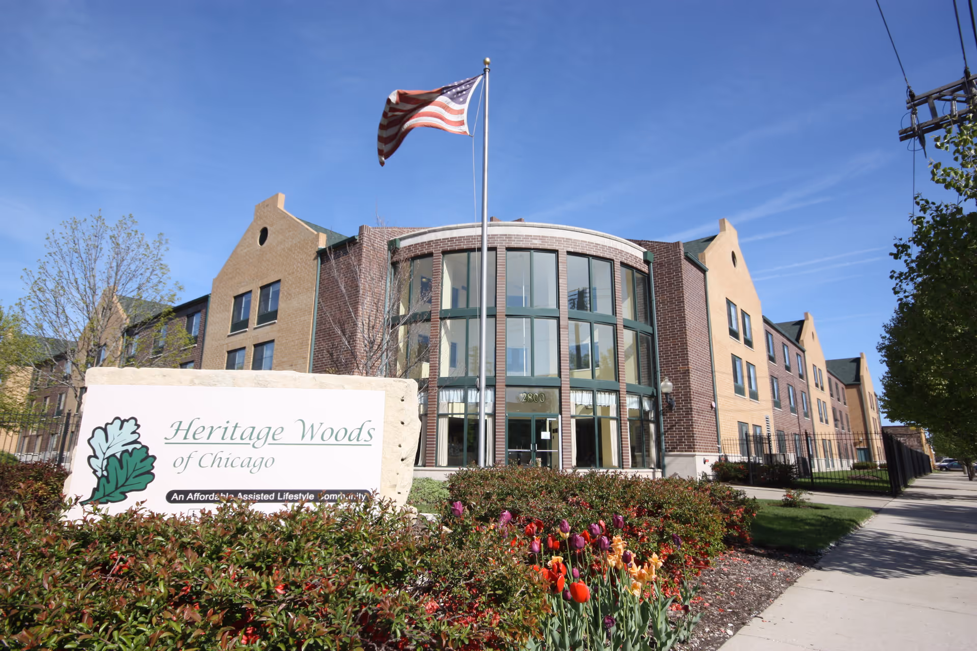 Exterior view of Heritage Woods of Chicago, a multi-story brick building with large windows and an American flag flying on a flagpole in front. There is a sign in the foreground with the facility's name surrounded by bushes and colorful flowers.