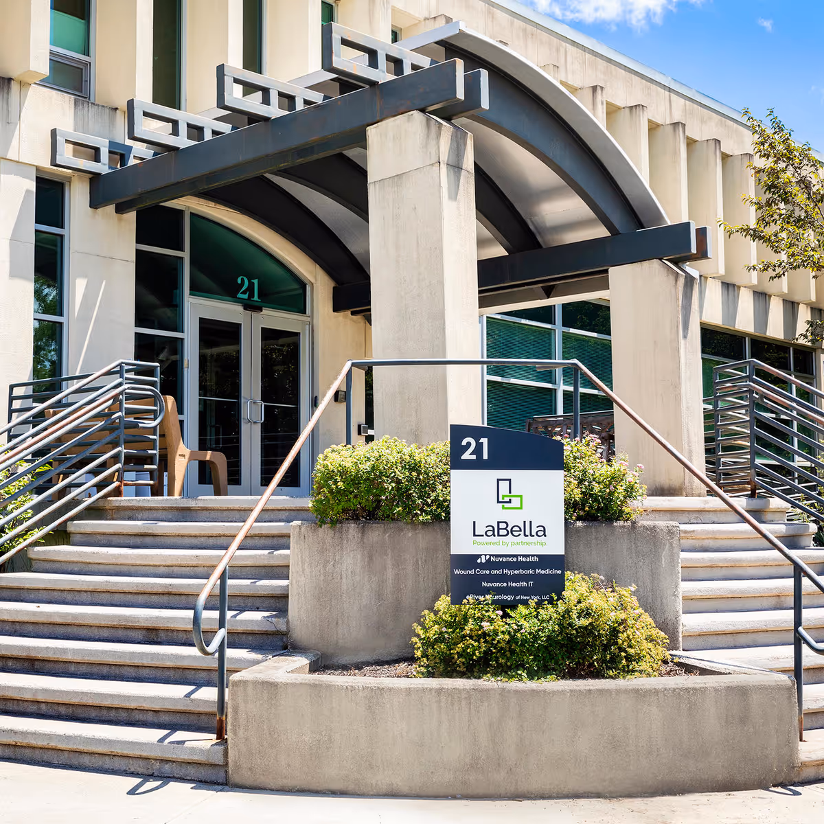 Front exterior entrance of a modern medical building with stairs, ramps, and a sign.